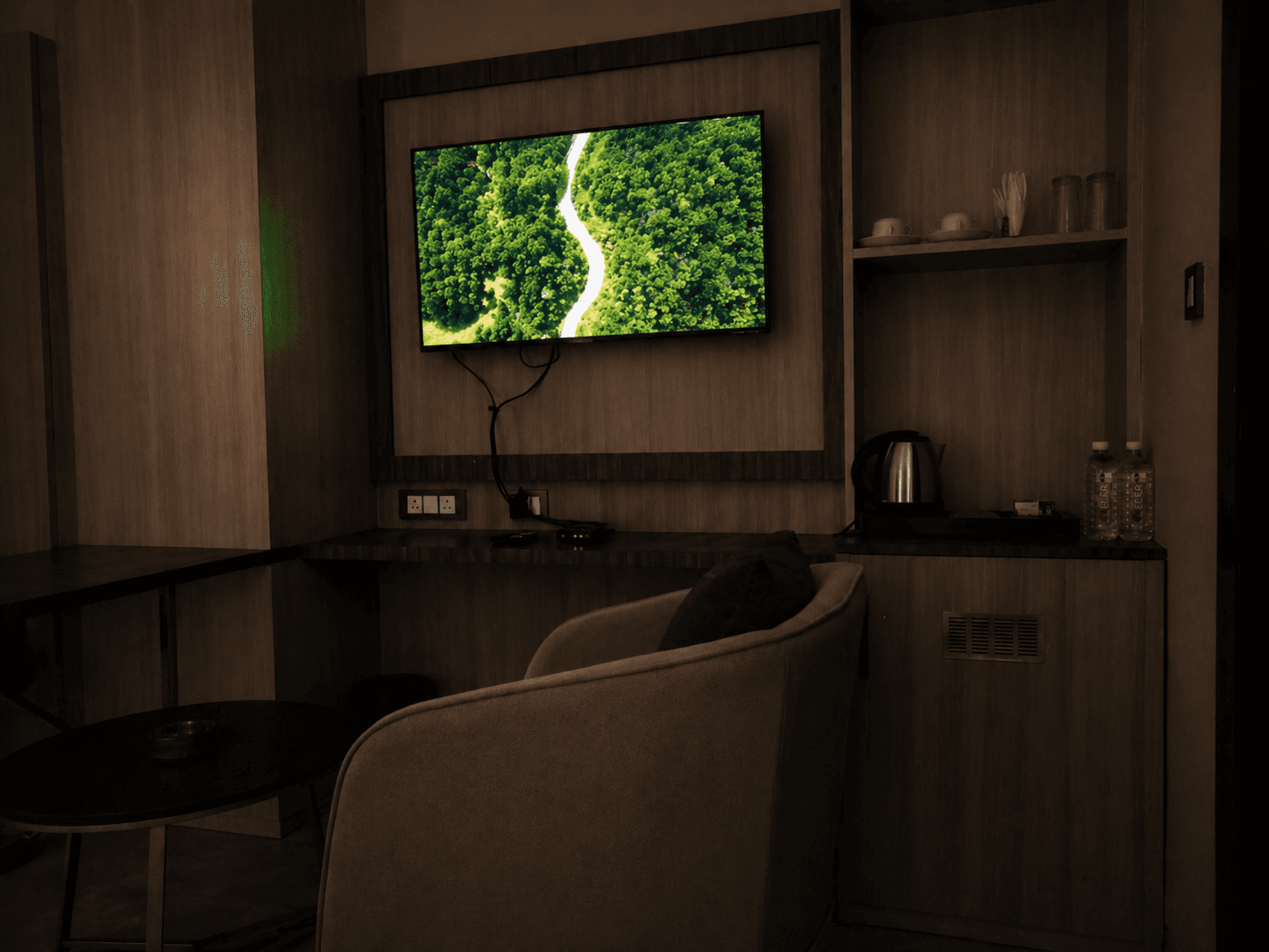 Television displaying a forest landscape above a grey armchair in a wood-paneled hotel room.