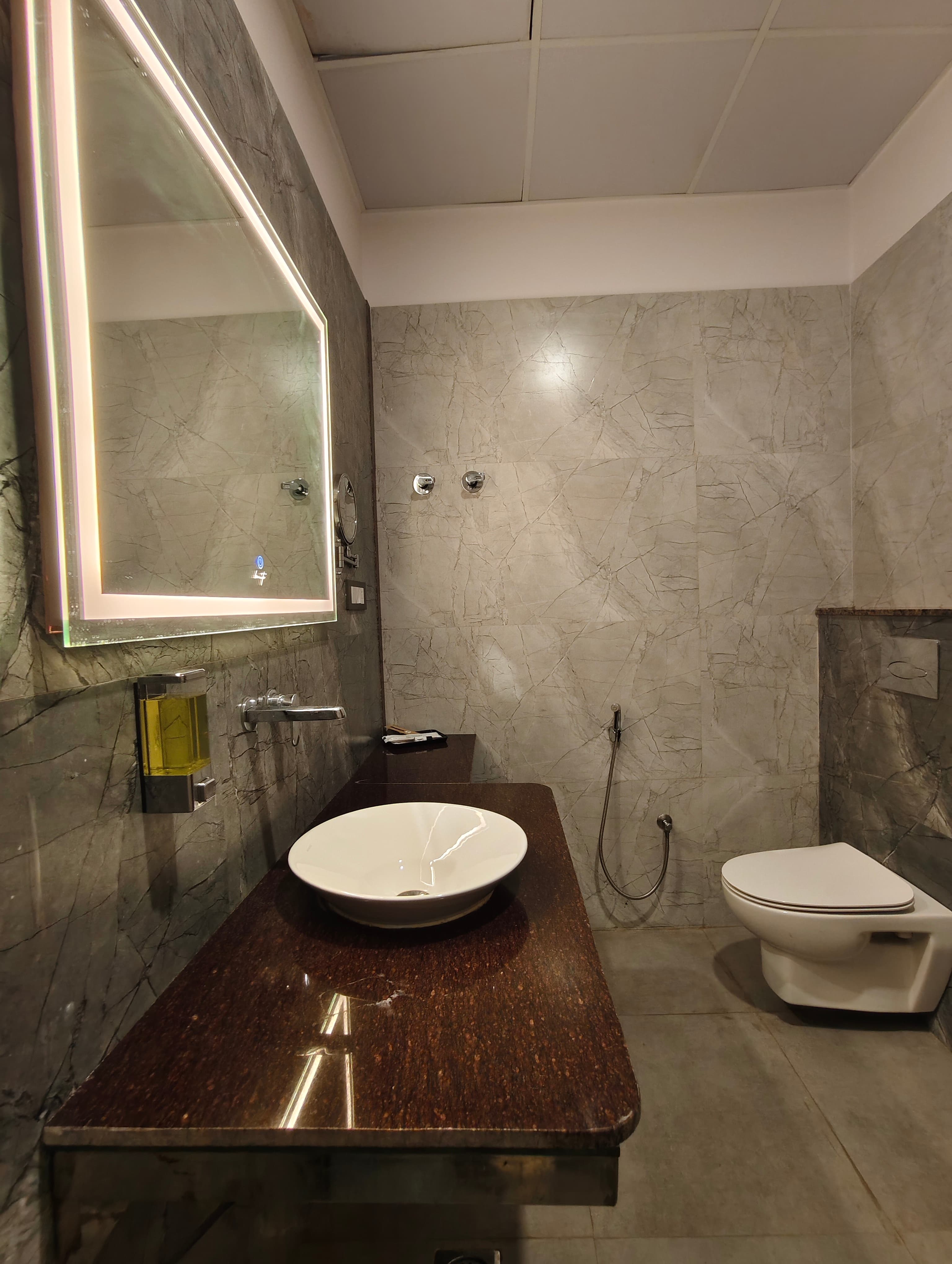 Modern bathroom with grey marble tiles, a backlit mirror, white vessel sink, and wall-hung toilet.