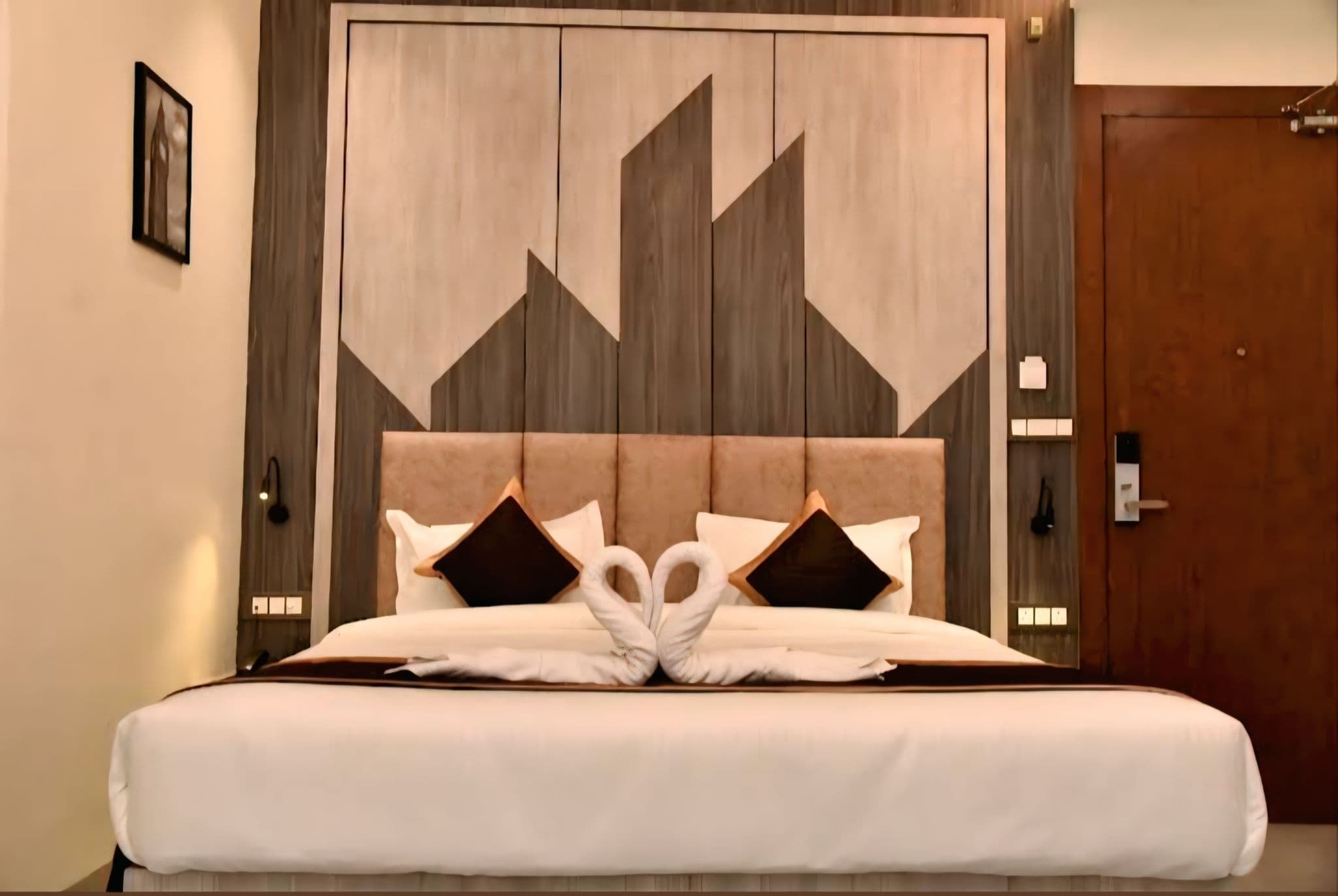 Two towel swans forming a heart on a hotel bed with a geometric wooden headboard.