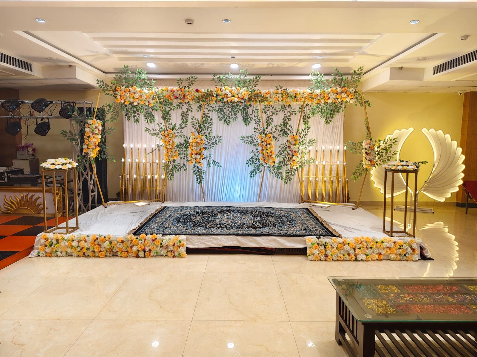 Elegant event stage with white backdrop, yellow floral arrangements, gold candle stands, and blue rug.
