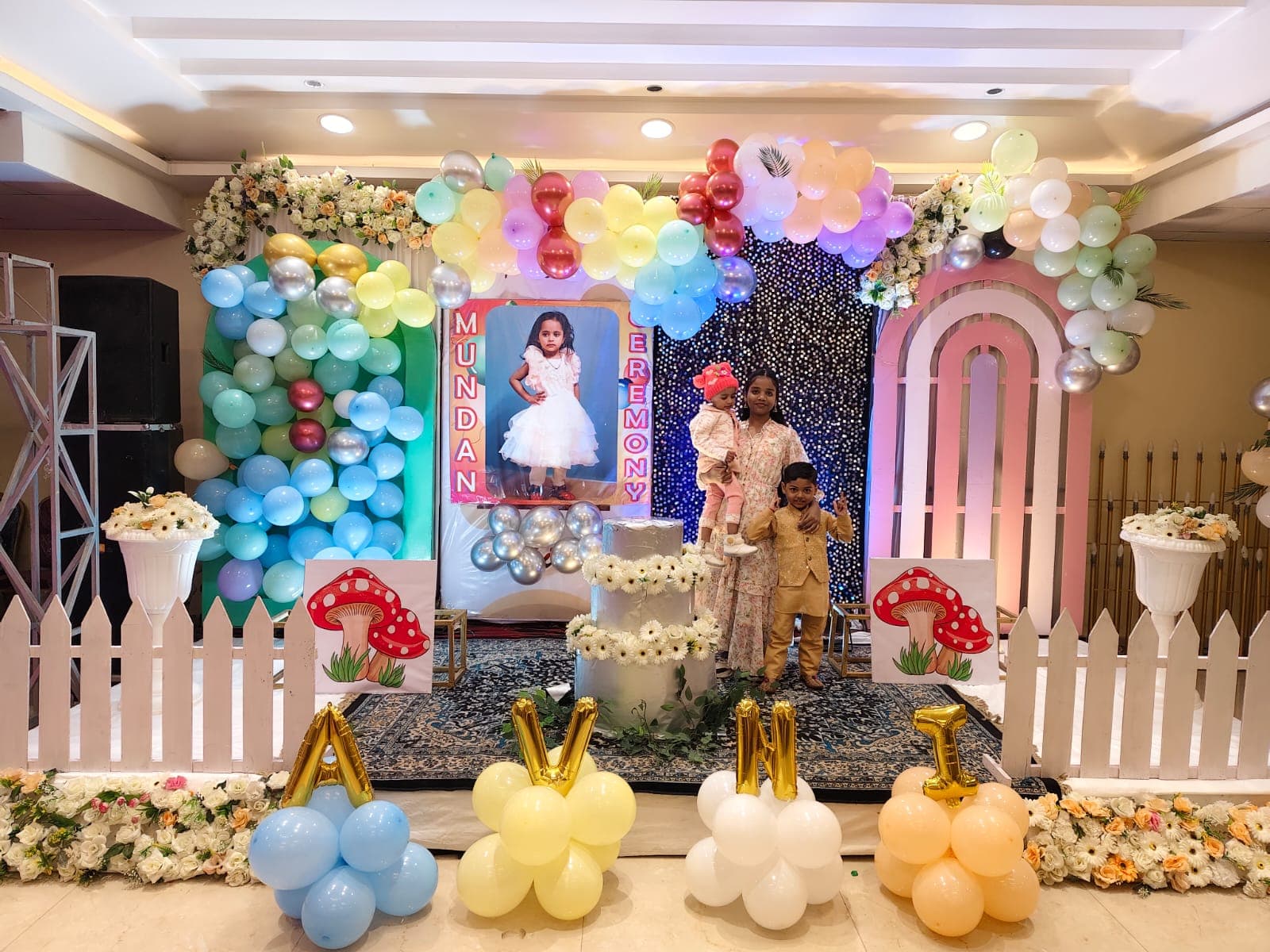 Family posing at a Mundan ceremony stage decorated with colorful balloons, flowers, and a poster.