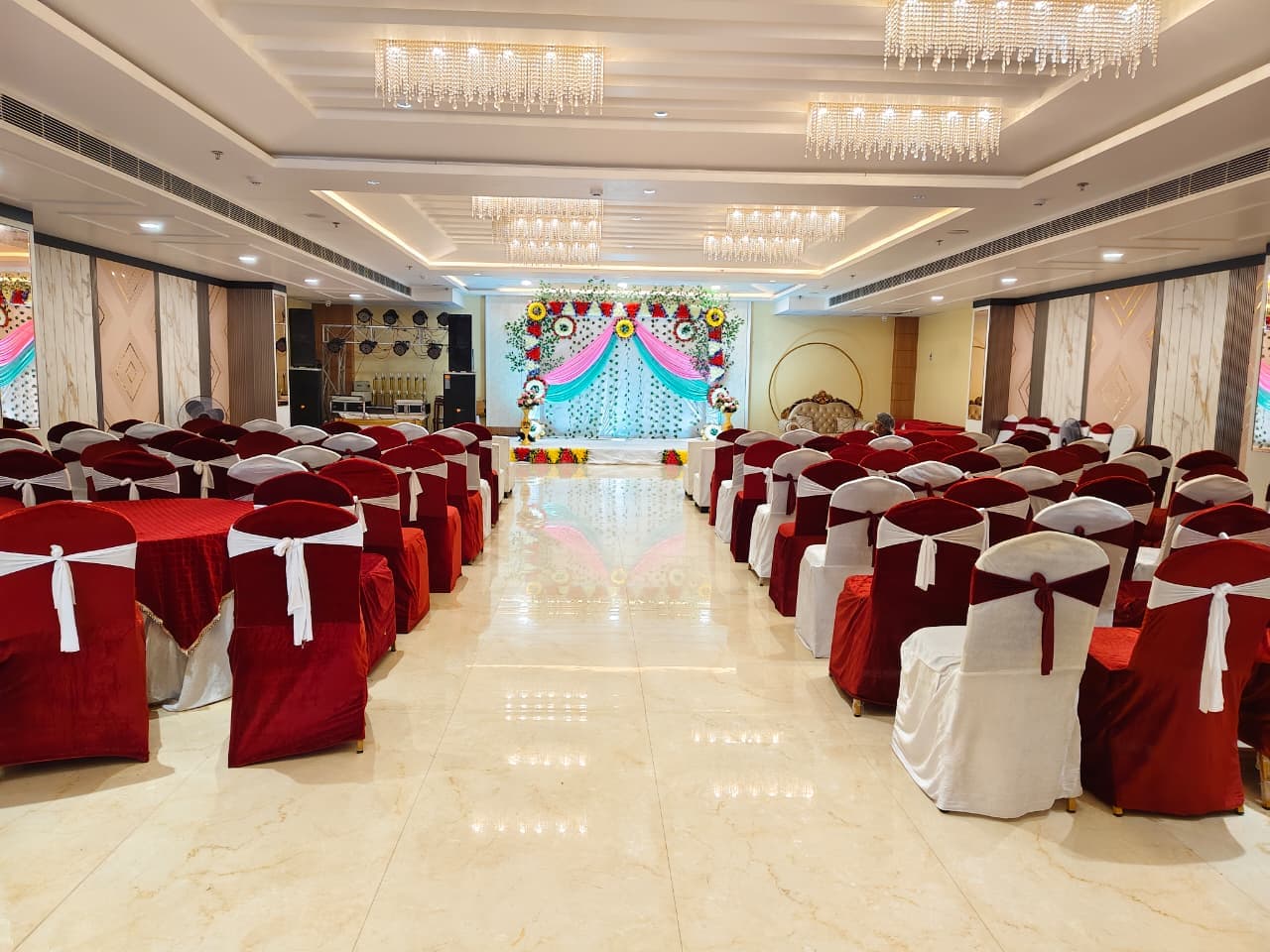 Elegant banquet hall featuring red chairs, crystal chandeliers, and a colorful floral stage backdrop.