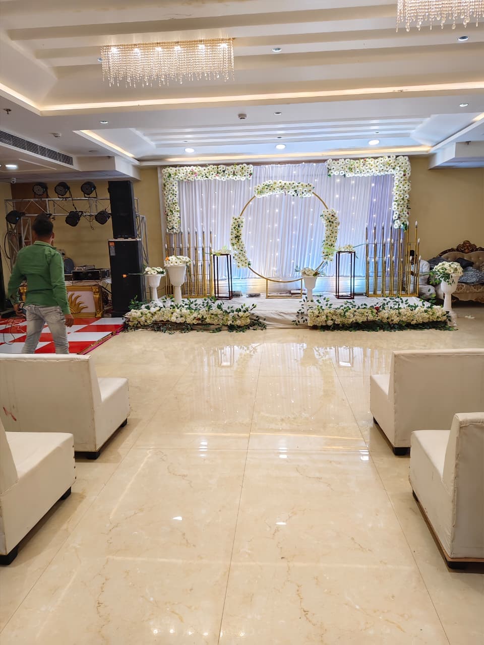 Elegant wedding stage with white floral decorations, gold circular frame, and white lounge seating.