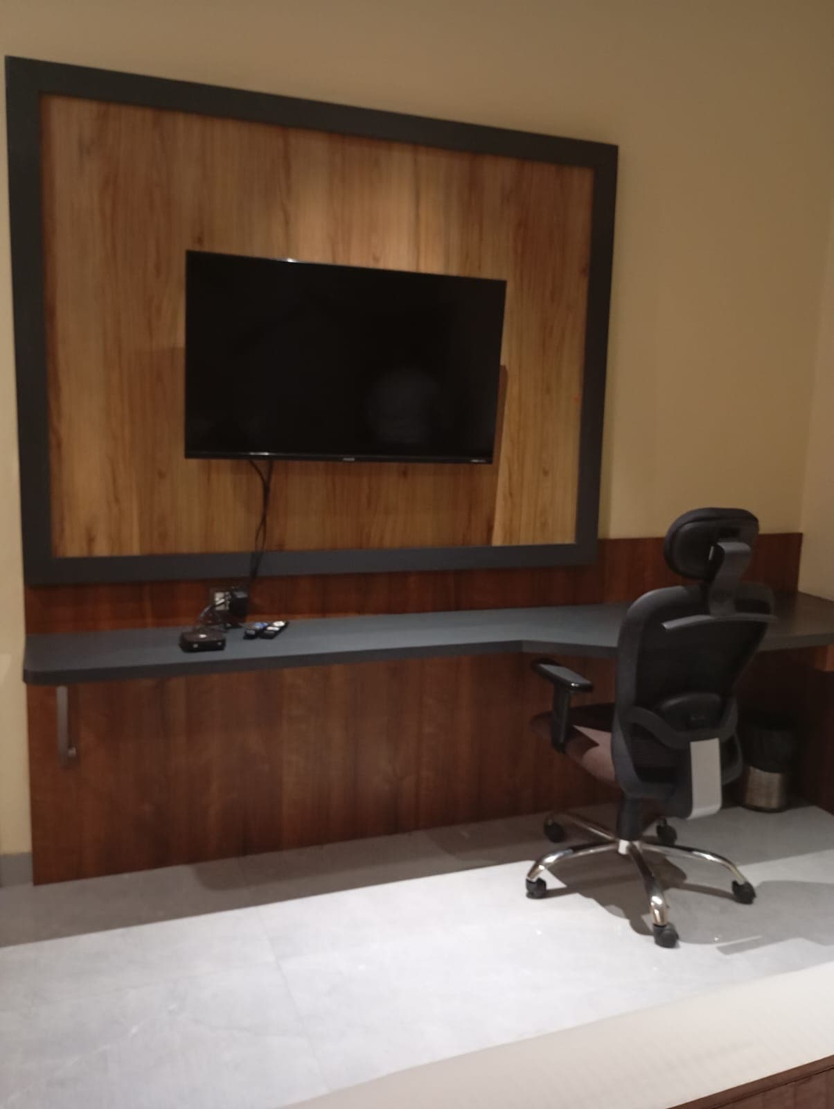 Wall-mounted TV on wood paneling above a grey desk with a black office chair.